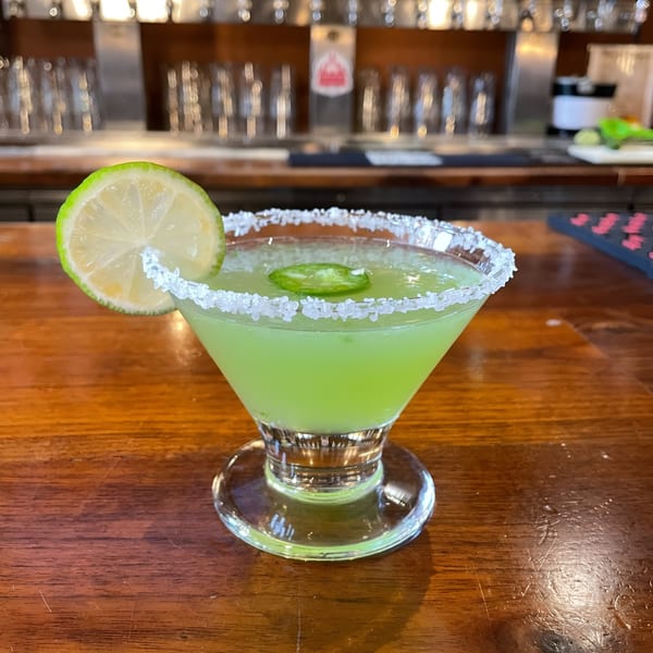 A vibrant green margarita with a salted rim, lime slice, and jalapeño on a wooden bar.