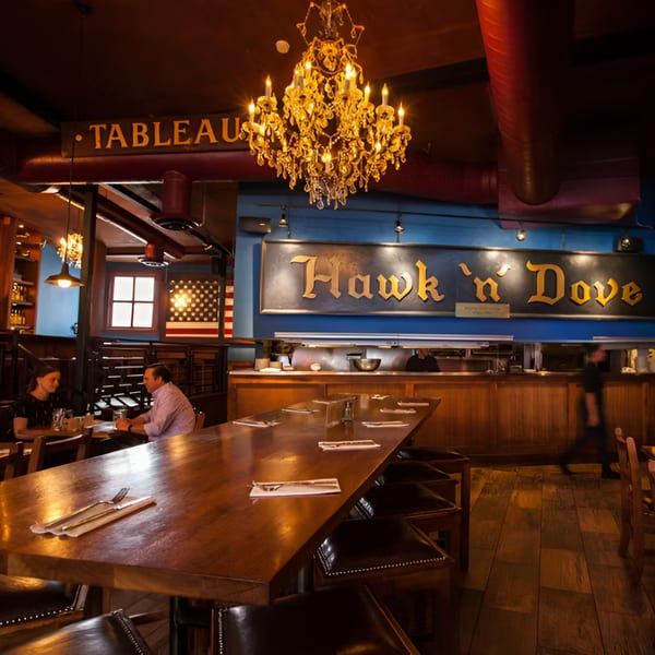 Interior of Hawk N Dove restaurant with a long wooden table, chandelier, and patrons dining.