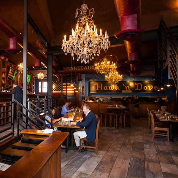 Interior of Hawk N Dove restaurant with patrons dining under ornate chandeliers and American flags.