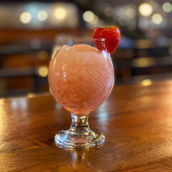 A frosty pink strawberry daiquiri with a fresh strawberry garnish sits on a wooden bar.
